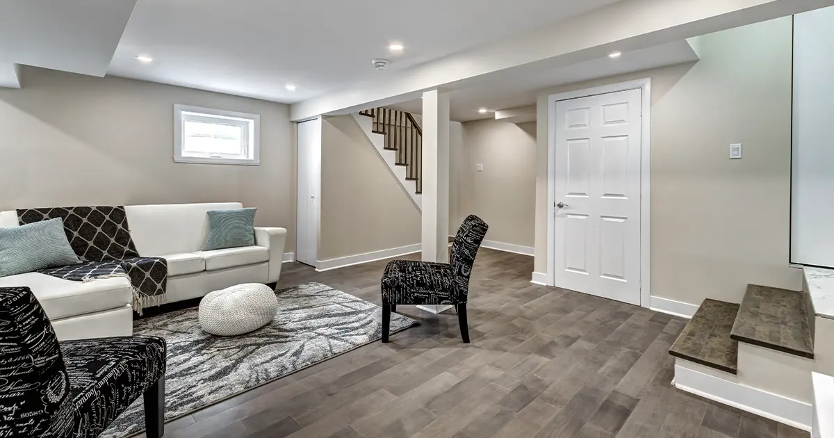 Modern finished basement lounge with white couch, wood flooring, recessed lights, and staircase with iron railing - featured