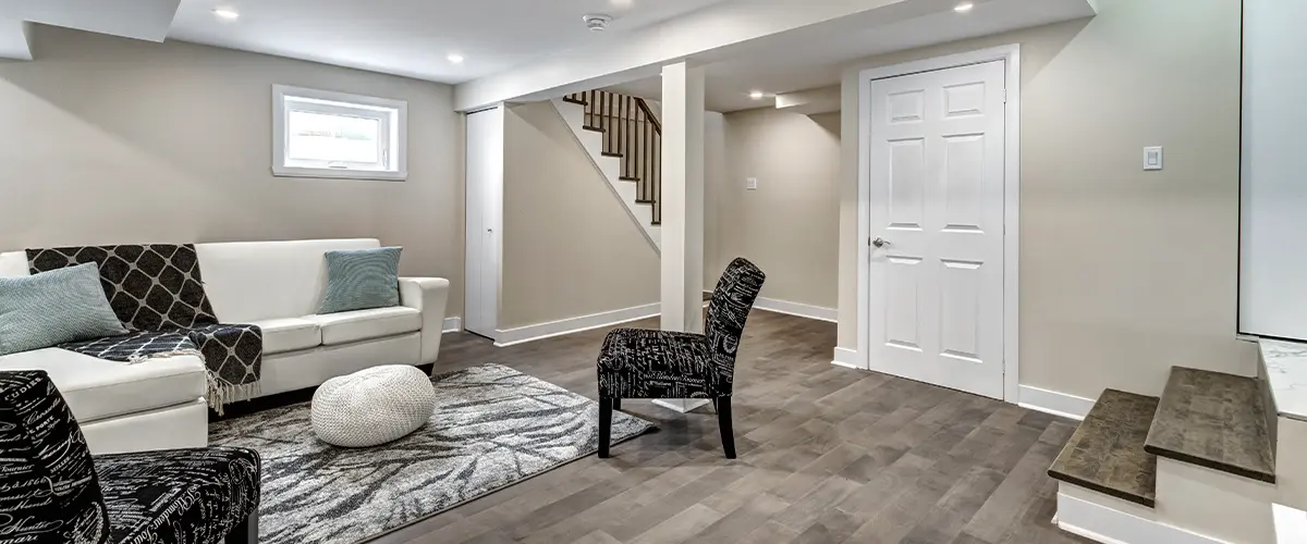 Modern finished basement lounge with white couch, wood flooring, recessed lights, and staircase with iron railing.