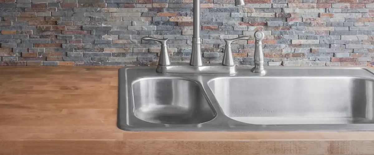Stainless steel double sink with wood countertop and stone backsplash.