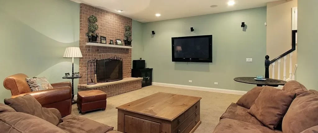 A traditional basement living room with a brick fireplace, mint green walls, large brown sofas, a wooden coffee table, and a wall-mounted TV.