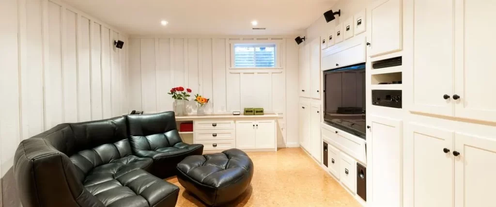 A cozy basement media room with white vertical paneling, a large built-in entertainment unit with storage cabinets, and a black leather sectional sofa and ottoman.