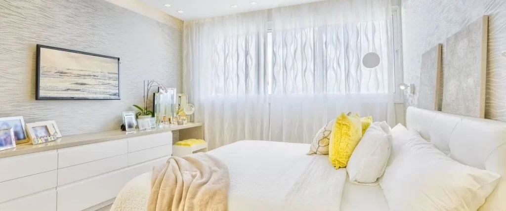 A modern basement bedroom with white furniture, sheer flowing white curtains over a large window, light textured wallpaper, and recessed ceiling lights.