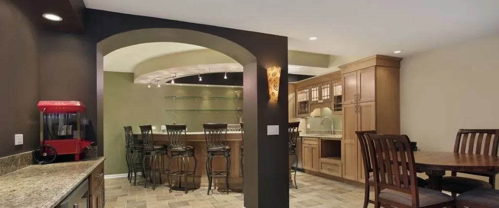 A fully finished basement recreation area with a wet bar, custom light wood cabinets, granite countertops, bar stools, a dining table, and a vintage red popcorn machine.