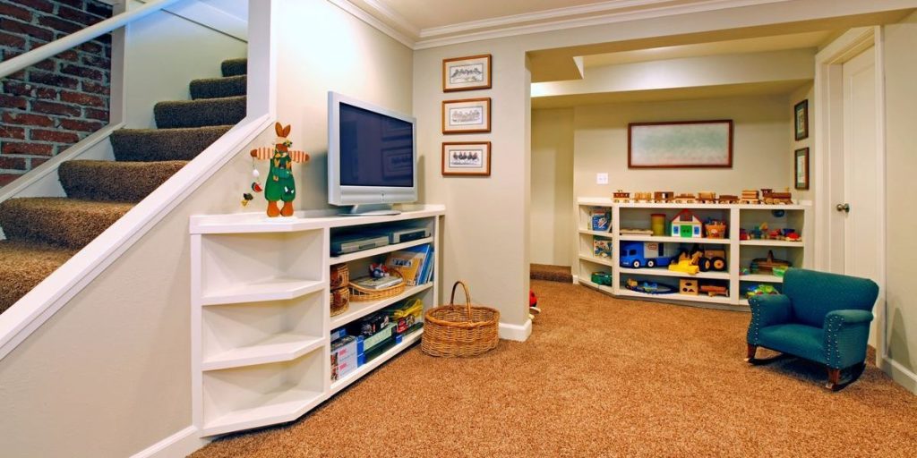A brightly lit, finished basement designed as a children's play area with thick brown carpet, built-in white shelving for toys, and a cozy rocking chair.