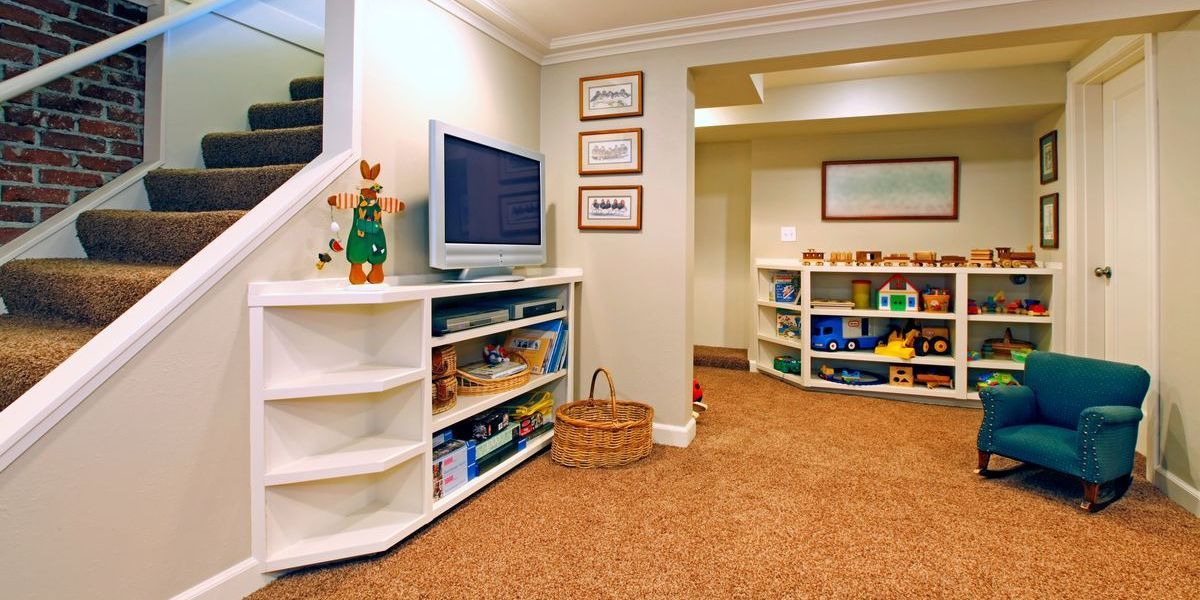 A brightly lit, finished basement designed as a children's play area with thick brown carpet, built-in white shelving for toys, and a cozy rocking chair.