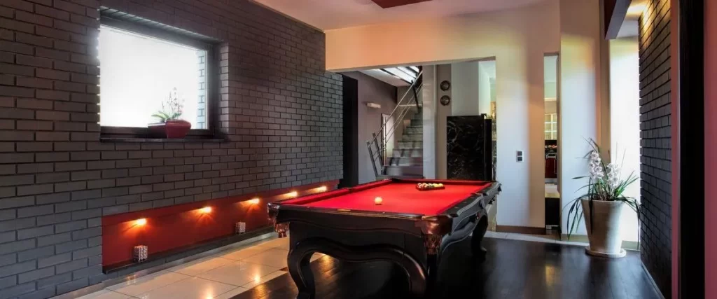 A luxurious, modern basement entertainment space featuring a red-felt billiard table, dark gray exposed brick wall, and stylish recessed lighting.
