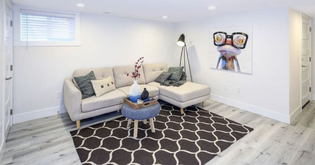 A bright, modern finished basement living area with white walls, a light beige sectional sofa, black geometric rug on gray wood-look laminate flooring, and a small window.