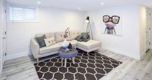 A bright, modern finished basement living area with white walls, a light beige sectional sofa, black geometric rug on gray wood-look laminate flooring, and a small window.
