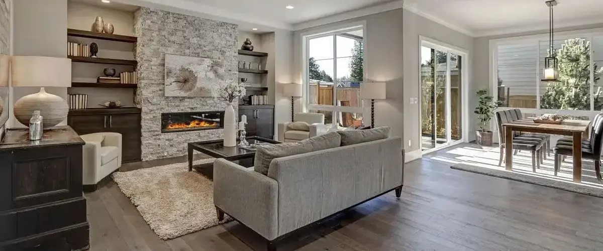 An open-concept living and dining space in a home addition with dark hardwood floors, a stone-accented fireplace wall, and large windows leading to an outdoor patio.