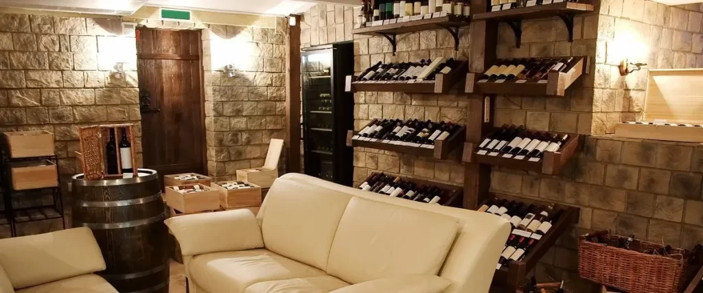 A rustic basement wine cellar and tasting room featuring rough stone walls, wooden wine racks, a small barrel table, and a comfortable white leather sofa.