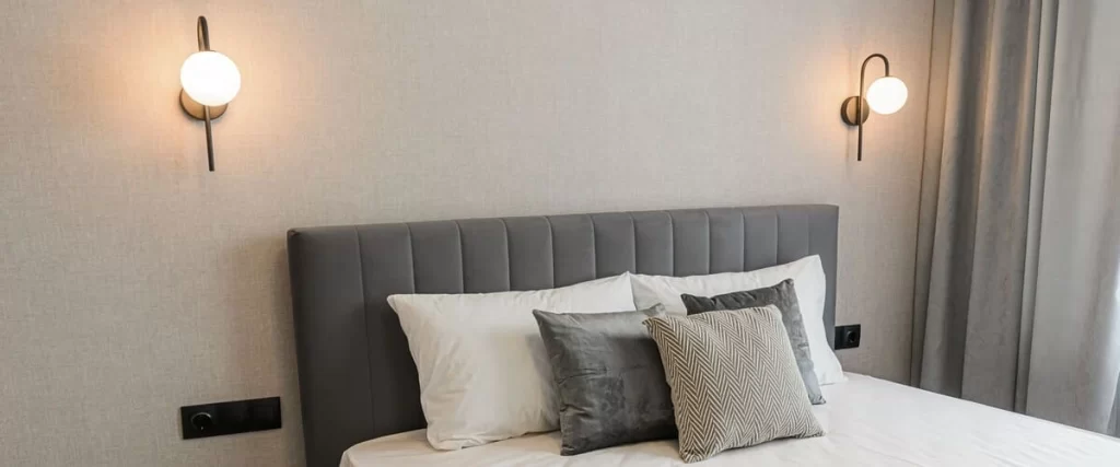 A close-up of a modern hotel-style bedroom featuring a gray tufted headboard, white pillows, and two elegant round white glass wall sconces mounted on a neutral textured wall.