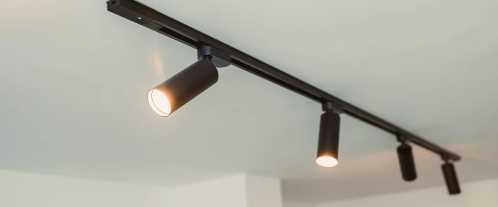 A close-up view of a sleek black track lighting system installed on a white ceiling with four adjustable cylindrical LED spotlights emitting a warm glow in a modern minimalist room.