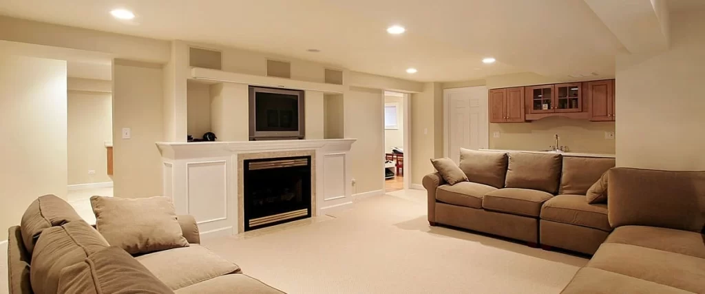 A spacious finished basement living area featuring warm recessed basement lighting, a white fireplace mantle with an integrated television, and a cozy tan sectional sofa near a small kitchenette bar.