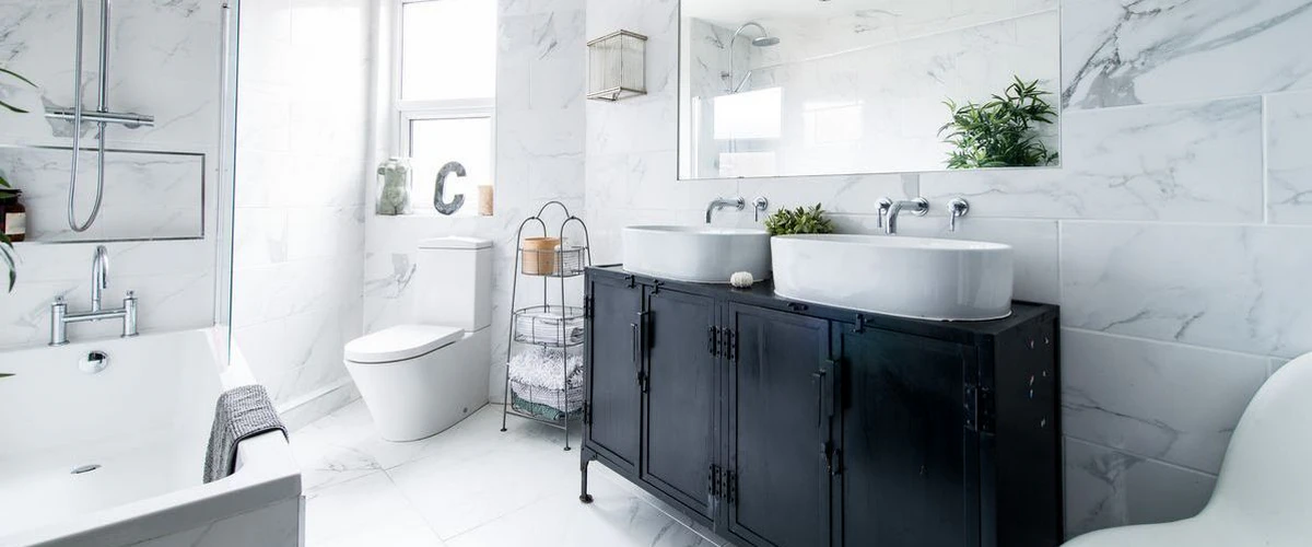 A bright, modern bathroom featuring white marble tile walls, a unique black vintage-style industrial metal vanity with double vessel sinks, and a large wall-mounted mirror reflecting a potted plant.