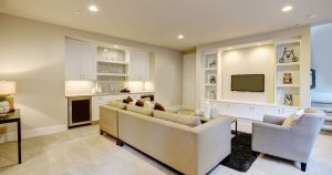 A modern finished basement living room showcasing layered lighting ideas for a basement including recessed ceiling fixtures, warm under-cabinet lights in a wet bar, and integrated display shelf lighting around a wall-mounted TV.