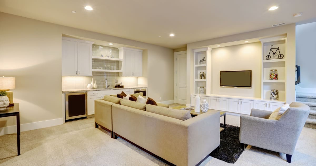 A modern finished basement living room showcasing layered lighting ideas for a basement including recessed ceiling fixtures, warm under-cabinet lights in a wet bar, and integrated display shelf lighting around a wall-mounted TV.