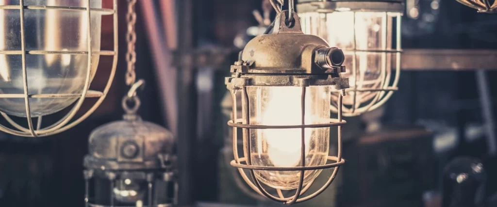 A close-up view of vintage industrial style lighting fixtures featuring weathered metal wire cages, glass casings, and warm glowing bulbs hanging in a dimly lit workshop.