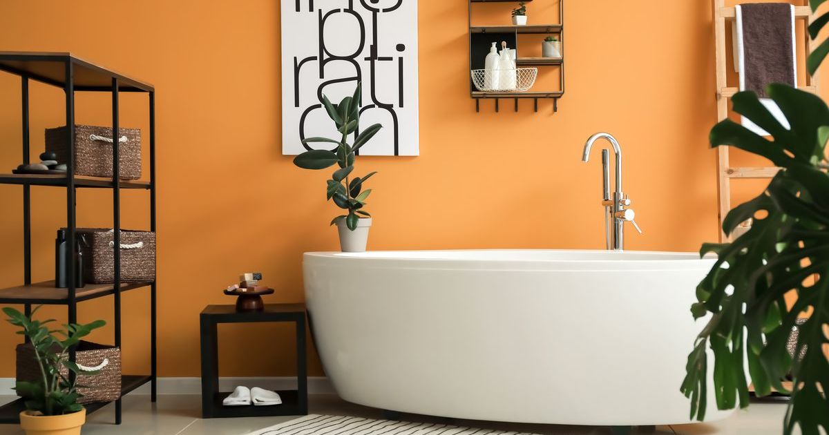 A luxury bathroom update by remodeling contractors in Glen Burnie, MD, highlighting a white soaking tub against a vibrant orange accent wall with black industrial shelving.