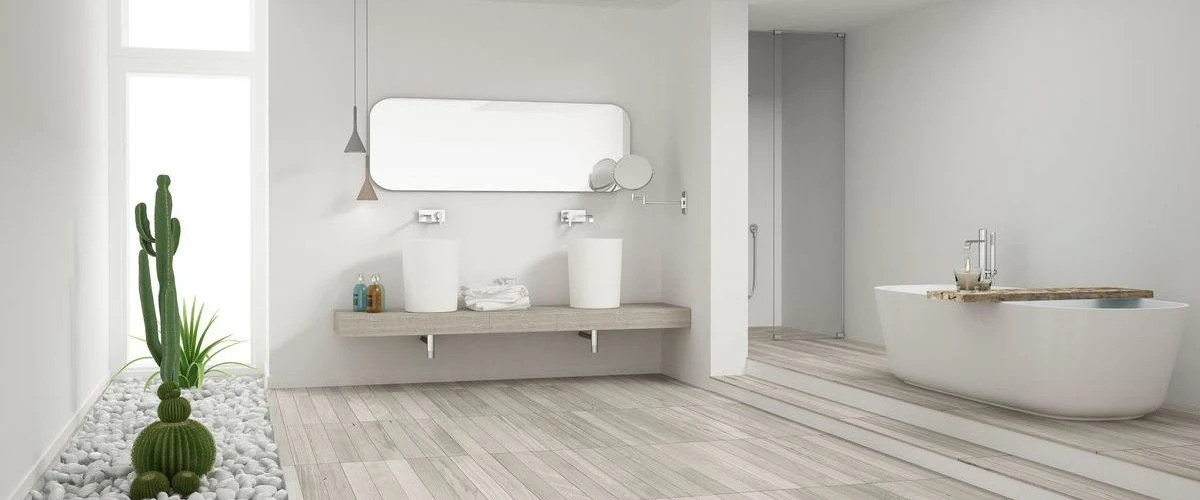 A wide-angle view of a minimalist, spacious bathroom with white walls, light wood flooring, a large freestanding bathtub, and a sleek double vanity setup.
