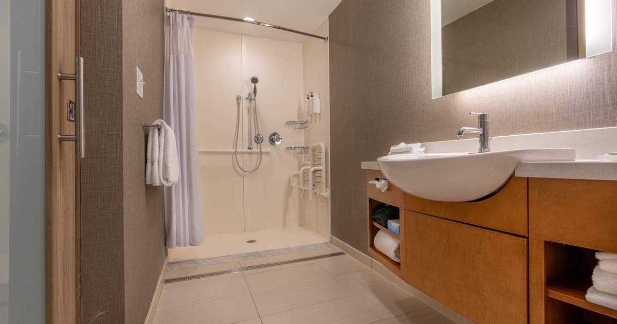 A comprehensive bathroom remodeling for seniors featuring a roll-in shower with a fold-down seat, multiple safety grab bars, and a modern wooden vanity with a backlit mirror.