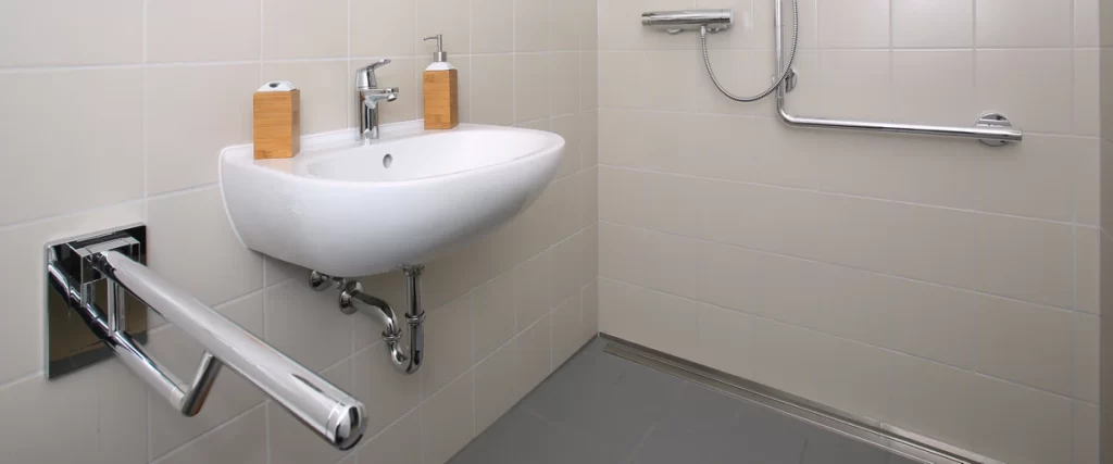 A minimalist bathroom design focused on accessibility for seniors, featuring a wall-hung sink for wheelchair clearance and a heavy-duty fold-down support rail.