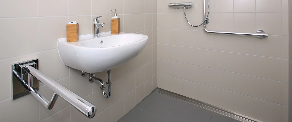 A minimalist bathroom design focused on accessibility for seniors, featuring a wall-hung sink for wheelchair clearance and a heavy-duty fold-down support rail.